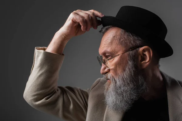 Side view of stylish senior man in beige jacket and eyeglasses adjusting derby hat posing on grey — Stock Photo