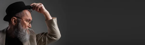 Side view of stylish senior man in beige jacket and eyeglasses adjusting derby hat posing on grey, banner — Stock Photo