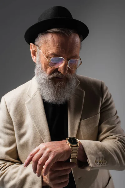 Elegante uomo anziano in giacca beige e cappello derby guardando orologio da polso su grigio — Foto stock