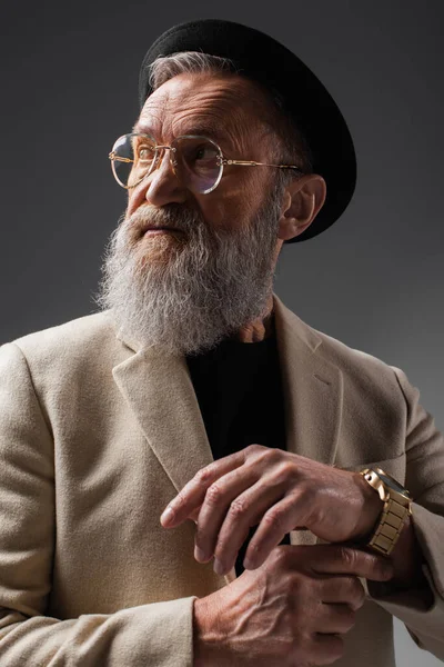 Elegante hombre senior en chaqueta beige y sombrero derby ajustando reloj de pulsera en gris - foto de stock