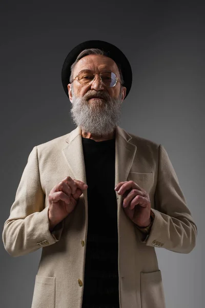 Stylish senior man in eyeglasses and derby hat adjusting woolen jacket on grey — Stock Photo