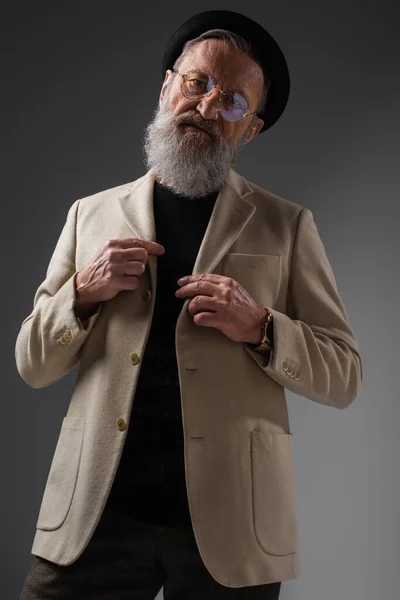 Elegant senior man in eyeglasses and derby hat adjusting woolen jacket on grey — Stock Photo