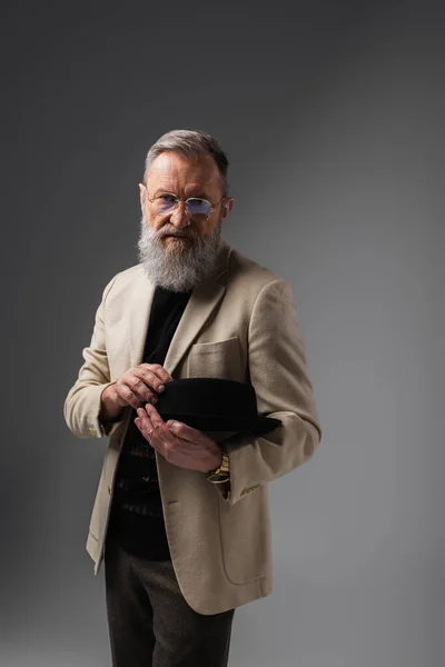 Stilvoller Senior in beiger Jacke und Brille mit Derbyhut auf grau — Stockfoto