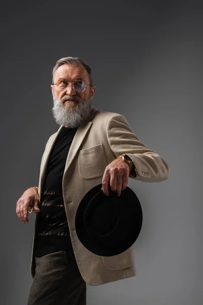 Elegant senior man in beige jacket and eyeglasses holding derby hat on grey — Stock Photo