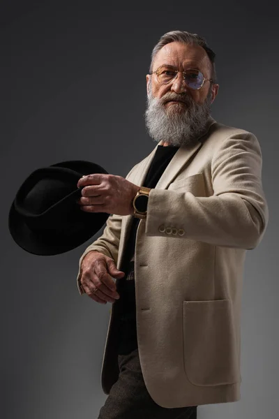 Bearded senior man in stylish jacket and eyeglasses posing with derby hat on grey — Stock Photo
