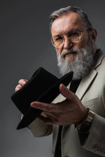 Bearded senior man in beige jacket and eyeglasses posing with derby hat on grey — Stock Photo