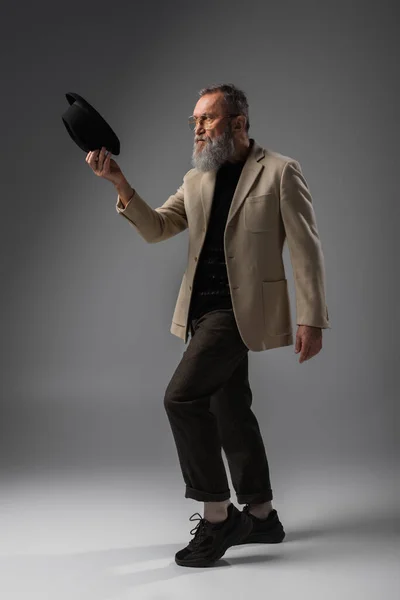 Full length of bearded senior man in beige jacket and eyeglasses posing with derby hat on grey — Stock Photo