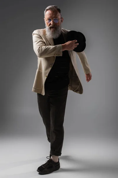 Full length of bearded senior man in stylish jacket and eyeglasses posing with derby hat on grey — Stock Photo