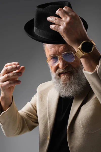 Elegant senior man in beige jacket and eyeglasses wearing derby hat on grey — Stock Photo