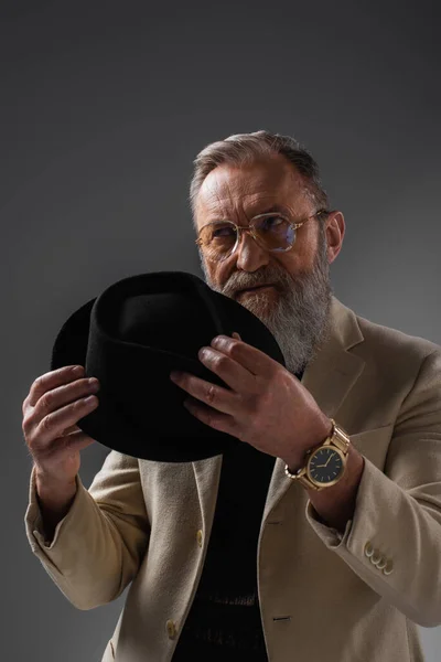 Елегантний старший чоловік у бежевій куртці та окулярах позує з дербі капелюхом на сірому — стокове фото