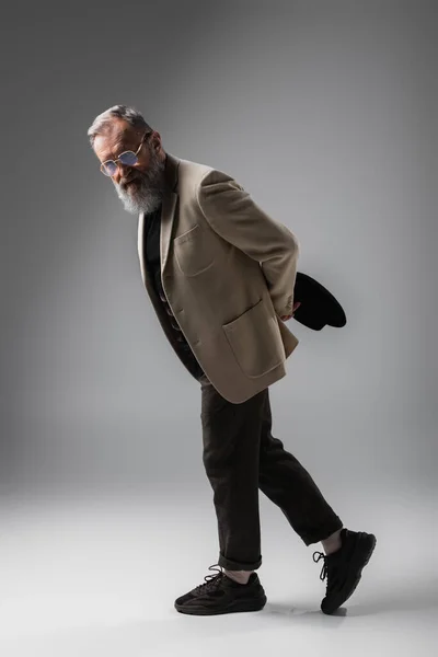 Full length of stylish senior man in beige blazer and eyeglasses posing with derby hat on grey — Stock Photo