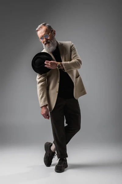 Full length of elegant senior man in beige blazer and eyeglasses posing with derby hat on grey — Stock Photo