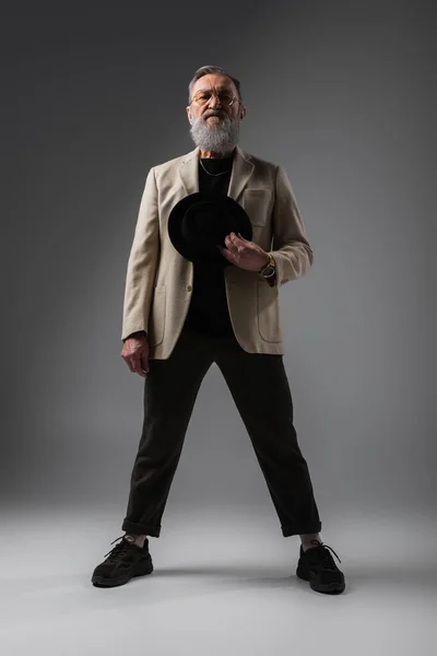 Longitud completa de elegante hombre senior en chaqueta beige y gafas de pie con sombrero derby en gris - foto de stock