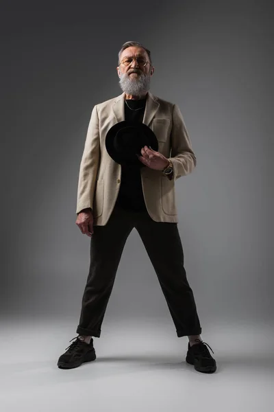 Full length of elegant senior man in beige jacket and eyeglasses standing with derby hat on grey — Stock Photo