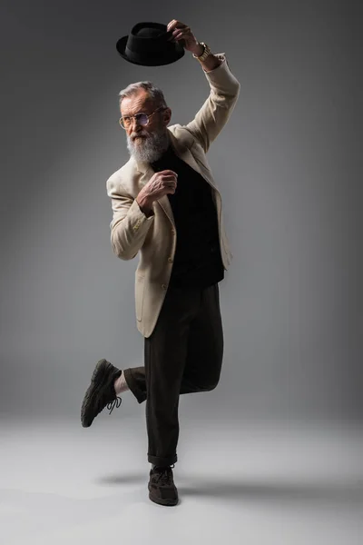 Full length of stylish senior man in beige jacket and eyeglasses posing with derby hat on grey — Stock Photo