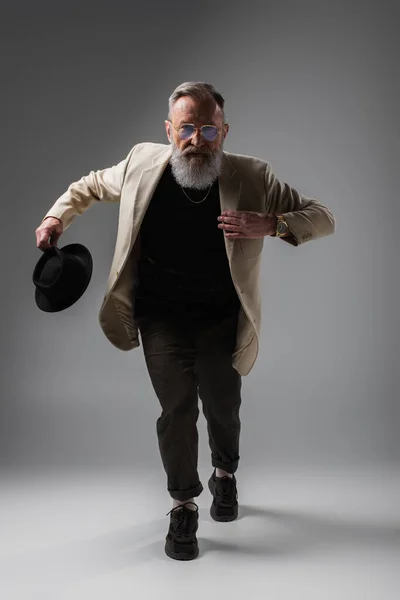 Full length of elegant senior man in beige jacket and eyeglasses posing with derby hat on grey — Stock Photo