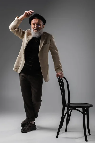 Comprimento total do homem sênior elegante na jaqueta bege e óculos posando com chapéu derby perto da cadeira no cinza — Fotografia de Stock