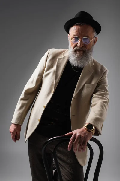Элегантный мужчина в бежевой куртке и очках позирует в шапке дерби рядом со стулом на сером — стоковое фото