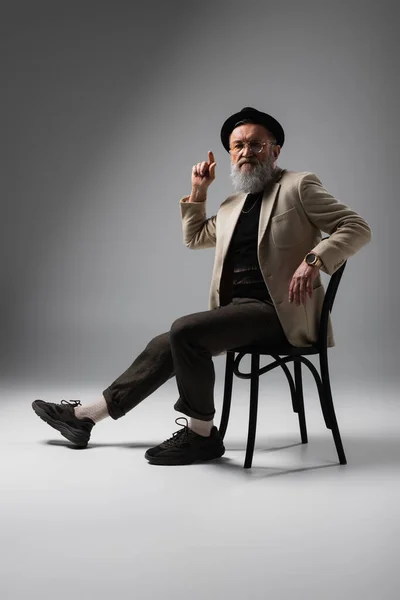 Full length of elegant senior man in beige jacket and derby hat sitting on wooden chair on grey — Stock Photo