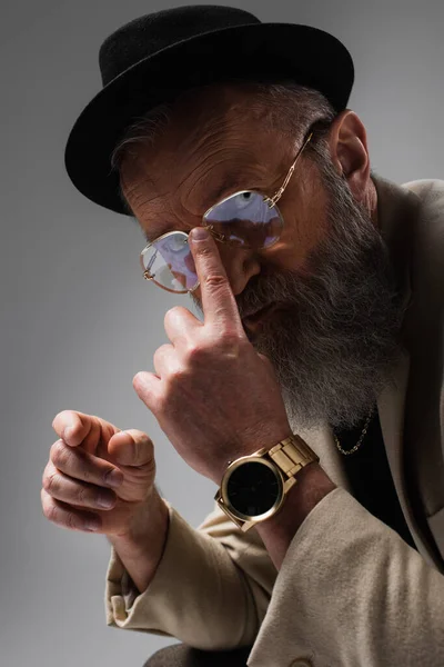 Retrato de hombre mayor barbudo en elegante chaqueta beige y sombrero derby ajustando las gafas con el dedo en gris - foto de stock