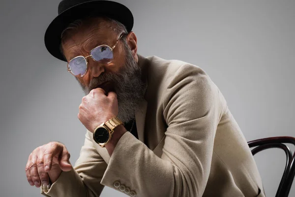 Portrait of bearded senior man in stylish beige jacket and derby hat looking at camera on grey — Stock Photo