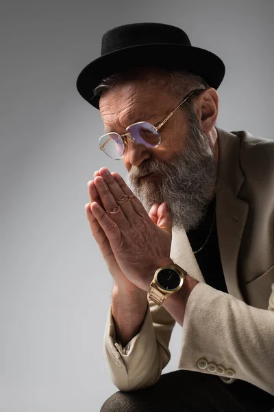 Portrait of bearded senior man in stylish beige jacket and derby hat with praying hands on grey — Stock Photo