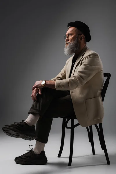 Бородатий старший чоловік у стильній бежевій куртці та капелюсі дербі сидить на дерев'яному стільці на сірому — стокове фото
