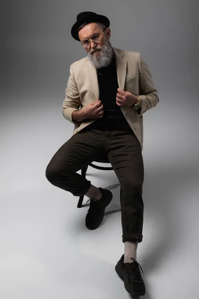 Full length of bearded senior man in stylish beige jacket and derby hat sitting on wooden chair on grey — Stock Photo