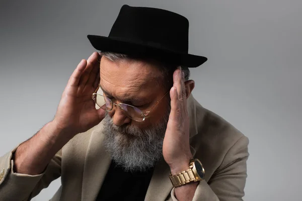 Uomo anziano barbuto in cappello derby e occhiali guardando giù su grigio — Foto stock