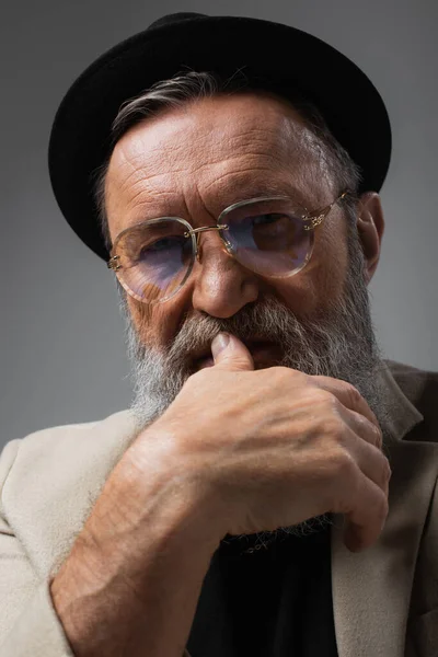 Ritratto di uomo anziano barbuto con occhiali e cappello da derby che guarda la macchina fotografica su grigio — Foto stock