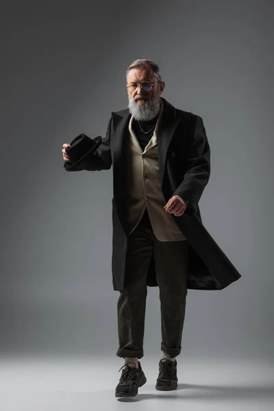 Full length of elegant senior man in stylish coat holding derby hat while walking on grey — Stock Photo