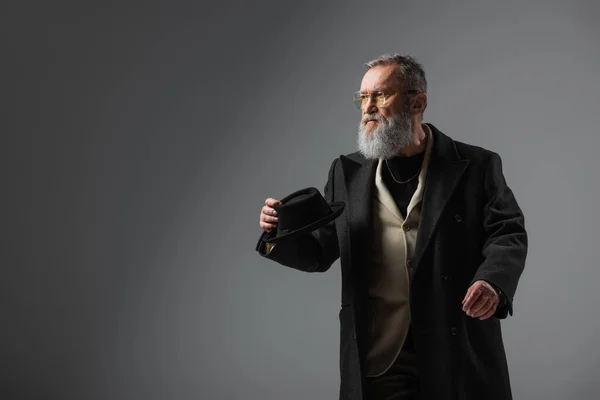 Elegant senior man in stylish coat and eyeglasses holding derby hat on grey — Stock Photo