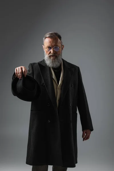 Stylish senior man in dark coat and eyeglasses holding derby hat on grey — Stock Photo