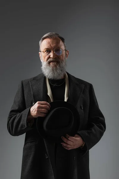 Бородатий старший чоловік у стильному пальто та окулярах тримає капелюх дербі на сірому — стокове фото
