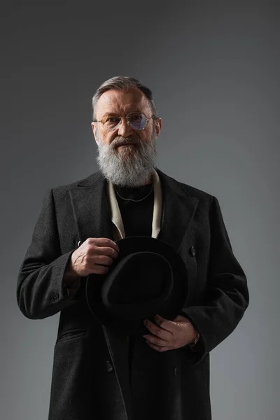 Homme âgé barbu en manteau élégant et lunettes tenant derby chapeau sur gris — Photo de stock