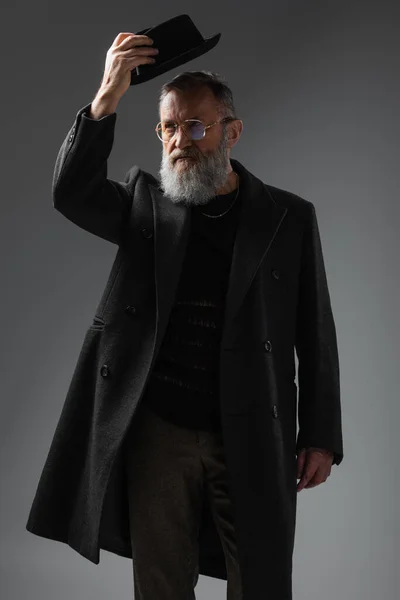 Élégant homme âgé en manteau élégant et lunettes de vue portant un chapeau derby sur gris — Photo de stock