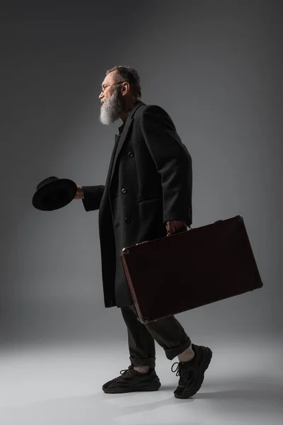 Full length of elegant senior man in stylish coat holding derby hat and suitcase on grey — Stock Photo