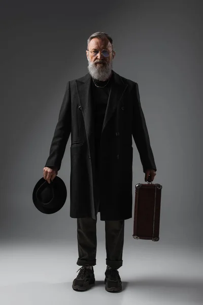 Full length of elegant senior man in coat holding derby hat and suitcase on grey — Stock Photo