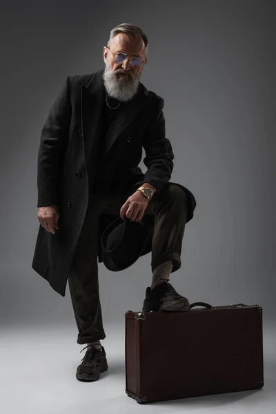 Full length of elegant senior man in stylish coat holding derby hat and putting leg on  suitcase on grey — Stock Photo