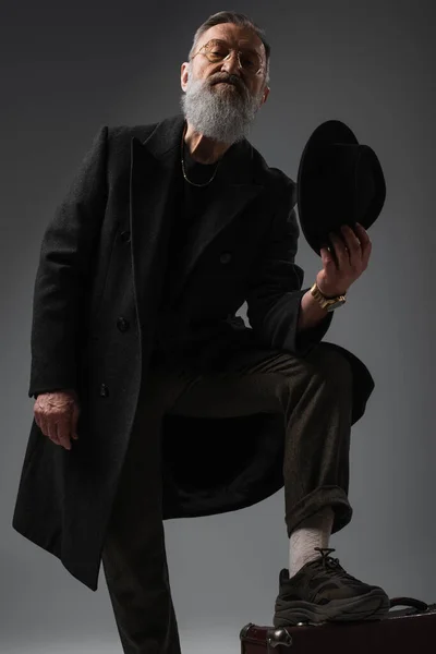 Bearded senior man in stylish coat holding derby hat and putting leg on suitcase on grey — Stock Photo