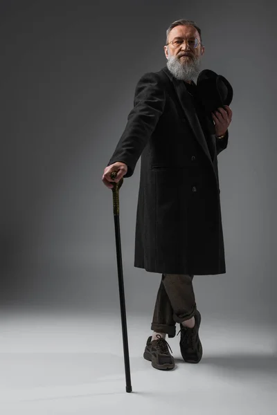 Comprimento total de homem sênior elegante em casaco elegante segurando chapéu derby e bengala andando em cinza — Fotografia de Stock