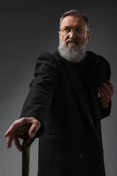 Elegant senior man in stylish coat holding derby hat and walking cane on grey — Stock Photo