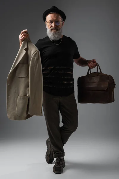 Full length of elegant senior man in derby hat holding leather bag and beige jacket on grey — Stock Photo