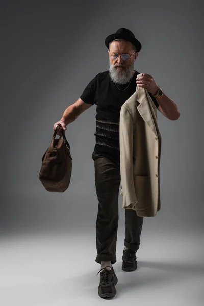 Full length of elegant senior man in derby hat holding leather bag and woolen jacket on grey — Stock Photo