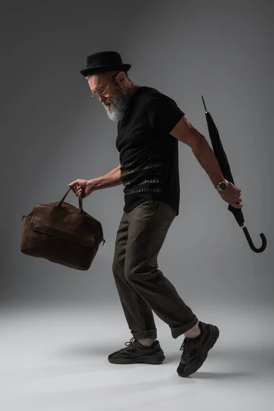 Toute la longueur de l'homme âgé élégant dans le chapeau derby tenant sac en cuir et parapluie sur gris — Photo de stock