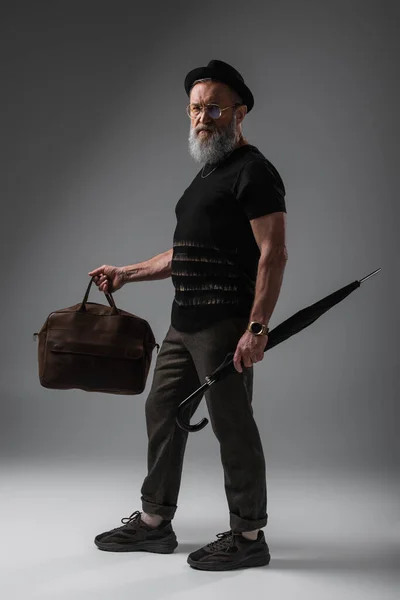 Полная длина бородатый мужчина в шляпе дерби дерби держа кожаную сумку и зонтик на сером — стоковое фото