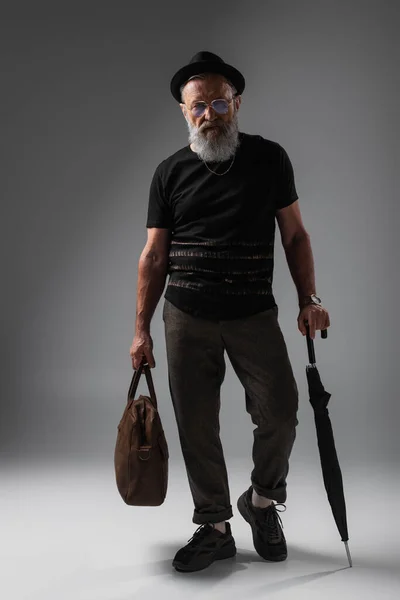 Full length of trendy senior man in derby hat holding leather bag and umbrella on grey — Stock Photo