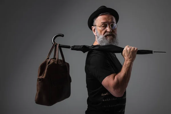 Stylish senior man in derby hat holding umbrella with leather bag on grey — Stock Photo