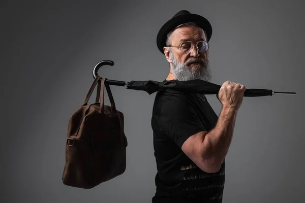 Homem sênior elegante em chapéu derby segurando guarda-chuva com saco de couro em cinza — Fotografia de Stock