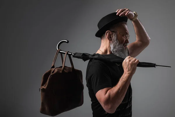 Side view of stylish senior man in derby hat holding umbrella with leather bag on grey — Stock Photo